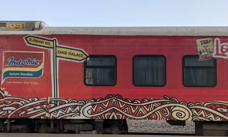 Red train car with Indomie noodle ad, colorful patterns, and street signs pointing to B.Gwari Rd. and Emir Palace.