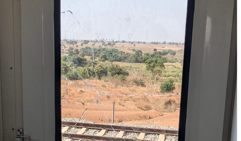 View through a cracked train window shows railway tracks and a rural landscape with scattered trees.