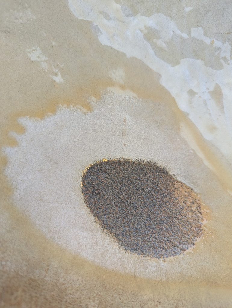 Close-up of a textured surface with an irregular dark spot surrounded by lighter, cloudy patterns.