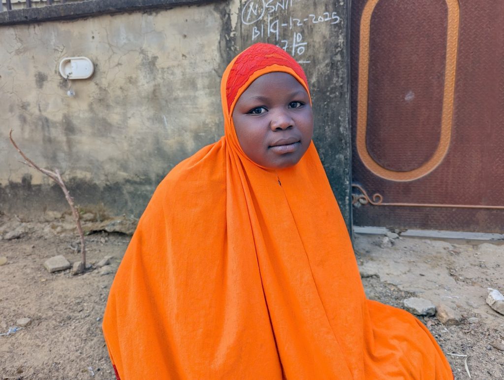 Young person in an orange hijab sits on the ground outdoors, with a wall and a gate in the background.