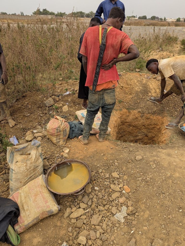 People working near a pit, with bags and a bowl of water on the ground, surrounded by dry vegetation.