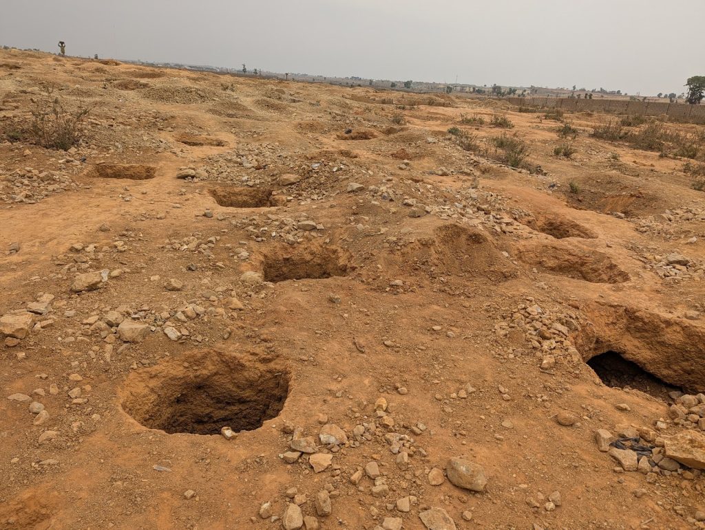 Dry, rocky landscape with several deep holes scattered across the surface under a cloudy sky. Sparse vegetation in the background.