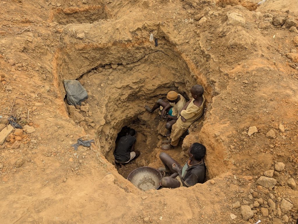 A group of four people sitting in a large hole dug in the ground, surrounded by loose dirt.
