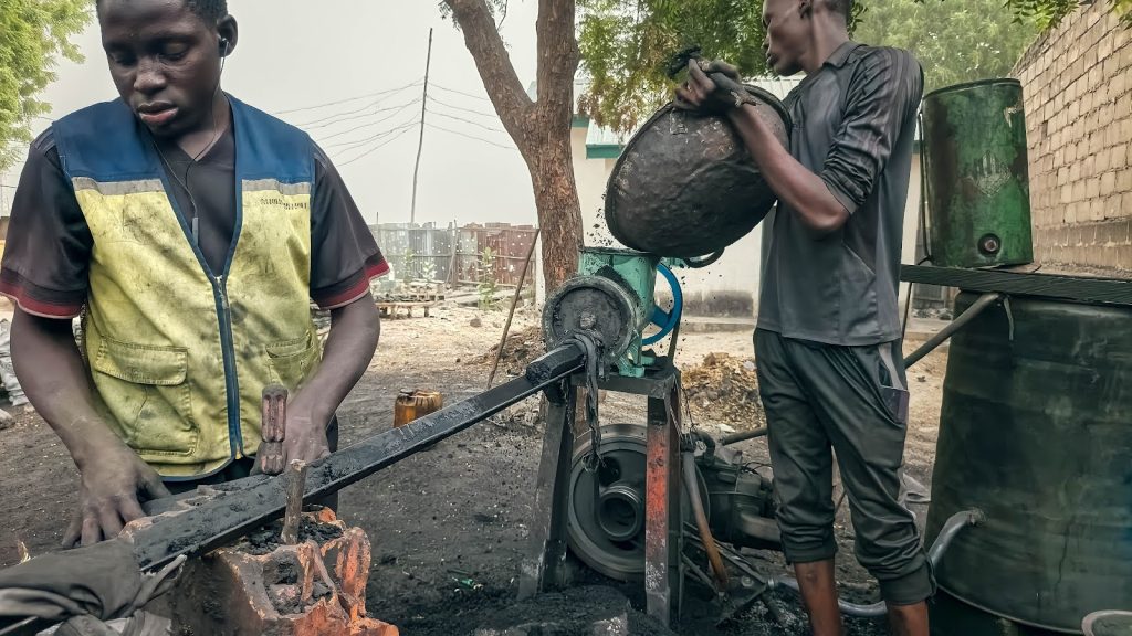 Two workers operate machinery outdoors; one adjusts controls while the other pours material into a hopper.