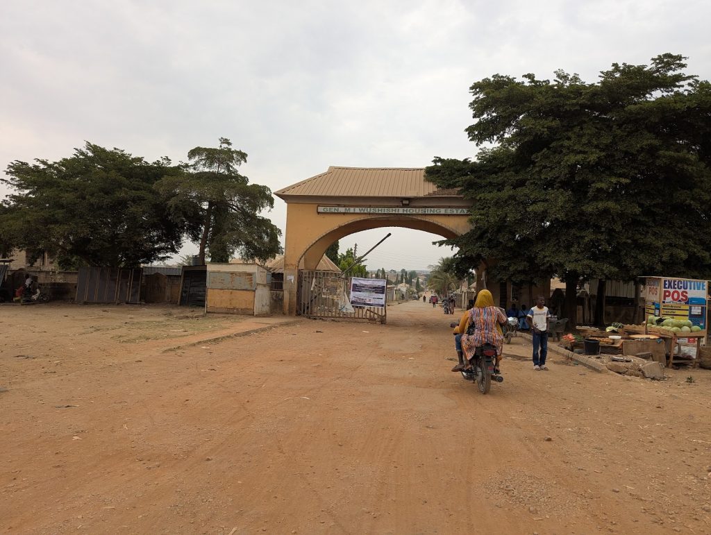A motorbike and pedestrians pass through a dusty archway entrance with surrounding trees and small shops.