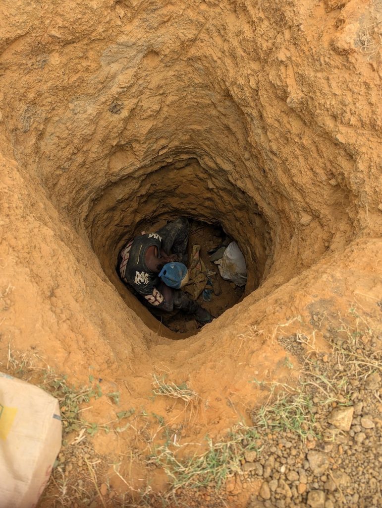 A person in a helmet and work gear is digging at the bottom of a deep earthen hole, surrounded by bags and dirt.