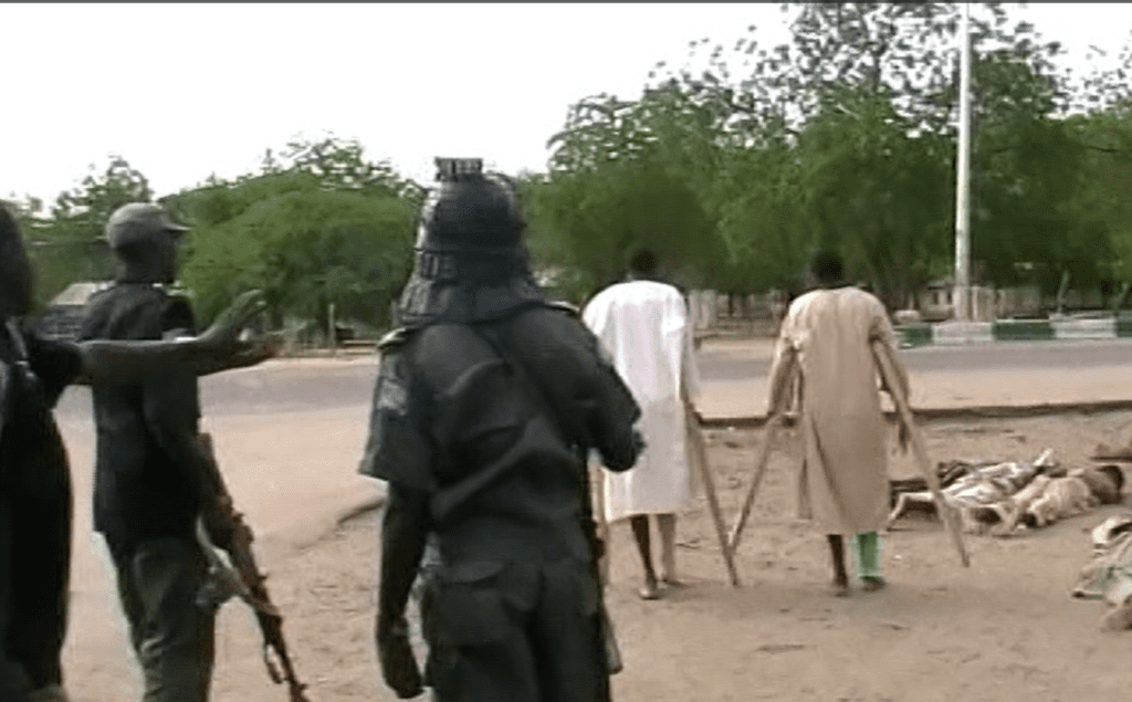 Armed personnel stand near two people on crutches, with more individuals lying on the ground in the background. Trees line the street.