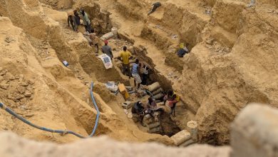 People working in a deep, narrow excavation site with exposed earth walls, carrying bags and using tools.