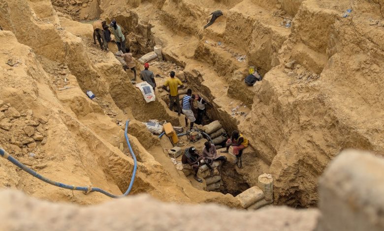 People working in a deep, narrow excavation site with exposed earth walls, carrying bags and using tools.