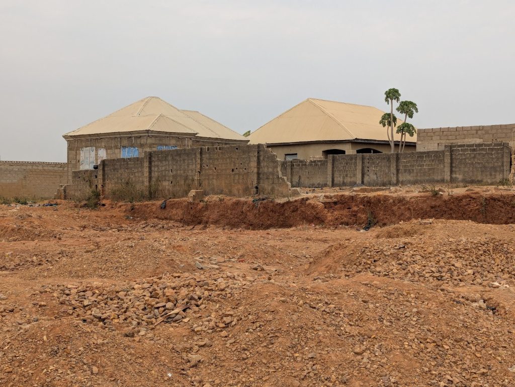 Houses with tin roofs behind a stone wall, surrounded by an empty, rocky, reddish-brown plot.