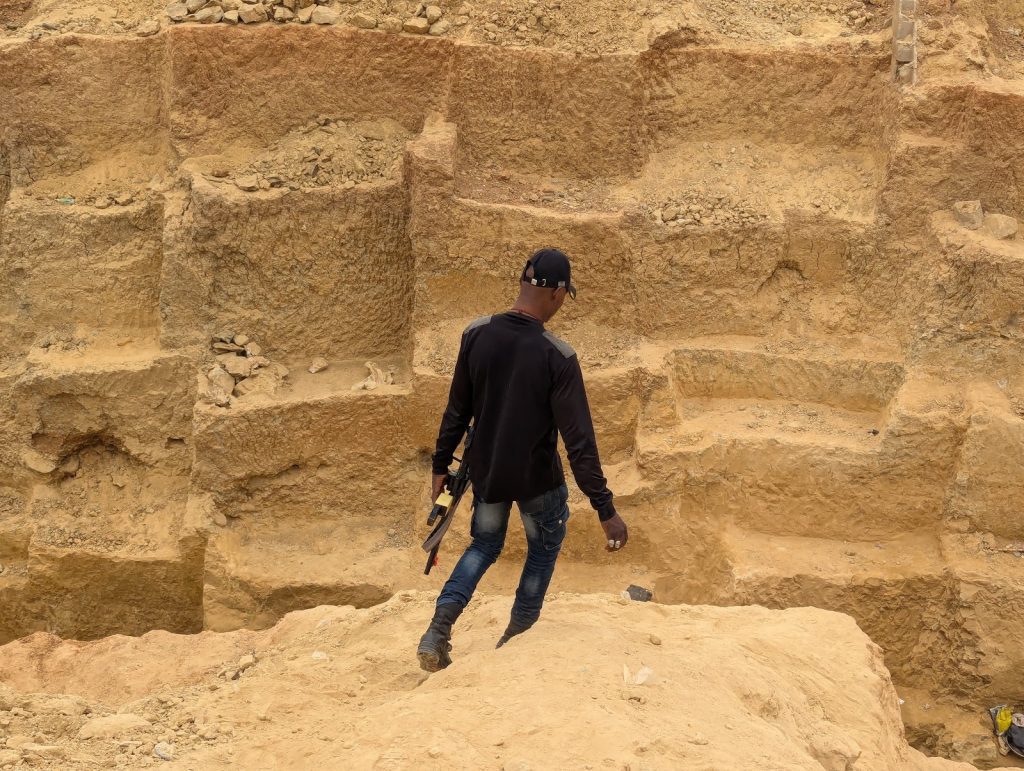 A person with a tool in hand walks down a stepped, rocky terrain, surrounded by earthy walls.