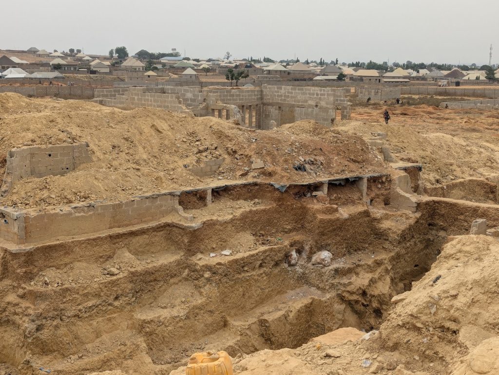 Excavated site with deep trenches and unfinished brick structures in a developing area, with houses visible in the background.