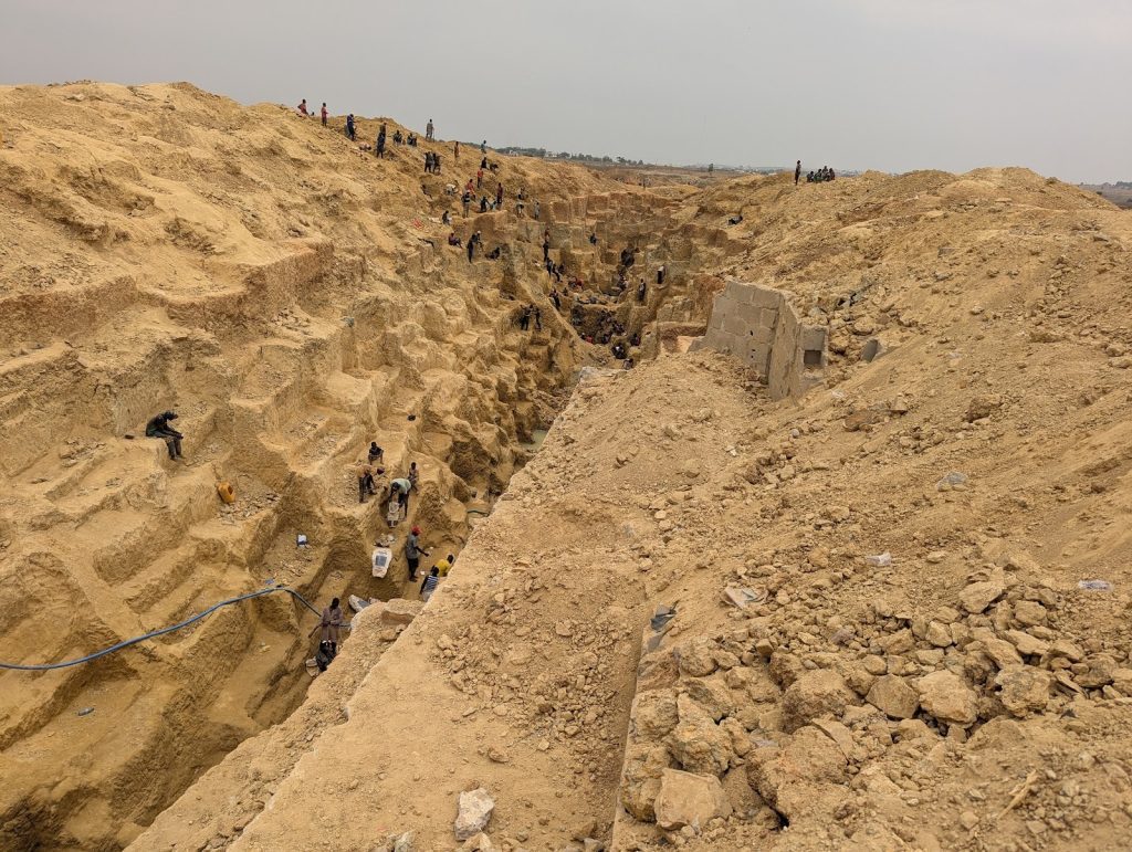 People working in a large, stepped sandy excavation site, scattered across different levels, with tools and equipment visible.