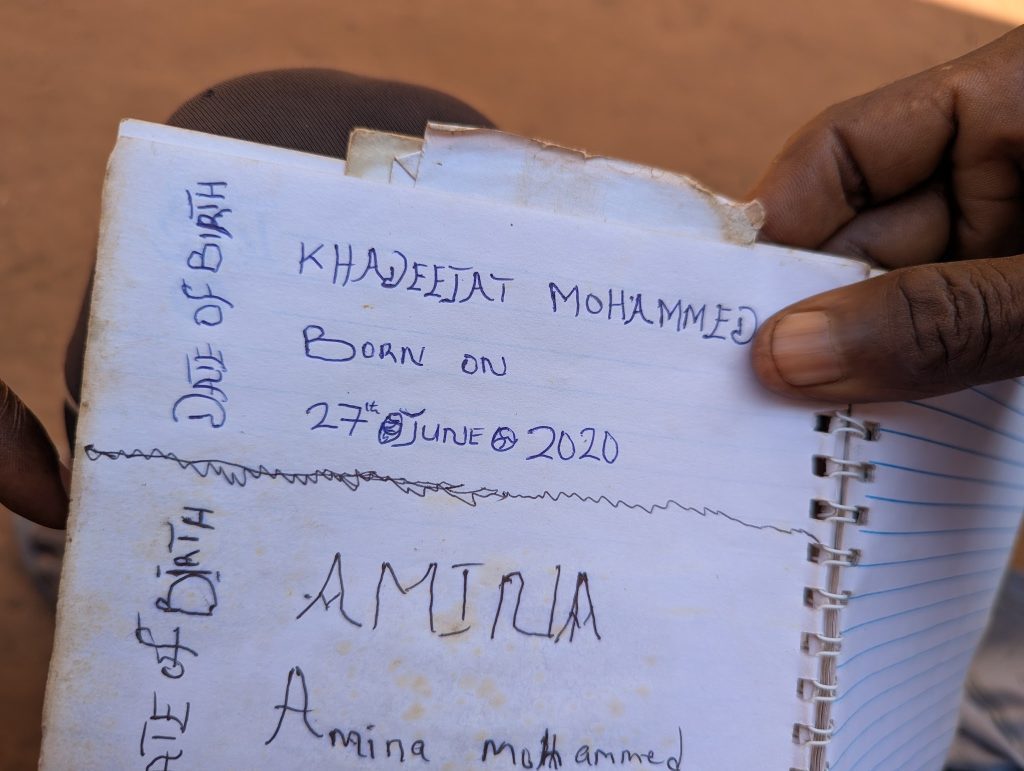 Close-up of a notebook page with handwritten text reading "Khadeejat Mohammed, born on 27th June 2020," held by a person's hand.