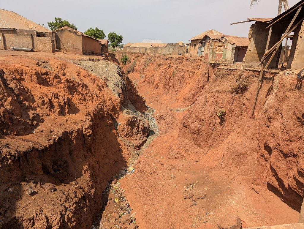Erosion forms a deep gully between houses with rust-colored soil and debris scattered, highlighting the impact on nearby structures.