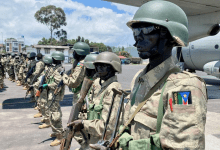 Soldiers in camouflage uniforms and helmets stand in formation near a military aircraft, with a South Sudan patch visible on a uniform.