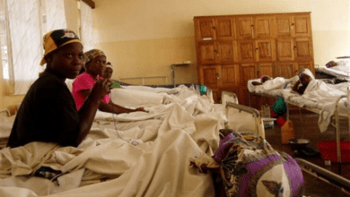 Hospital ward with several patients in beds, some sitting up, with wooden cabinets in the background.