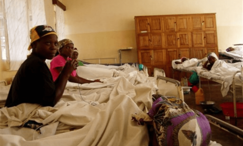 Hospital ward with several patients in beds, some sitting up, with wooden cabinets in the background.