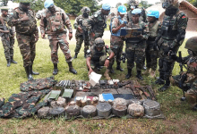 Soldiers in camouflage and UN helmets examine items laid out on the ground, discussing and taking notes.