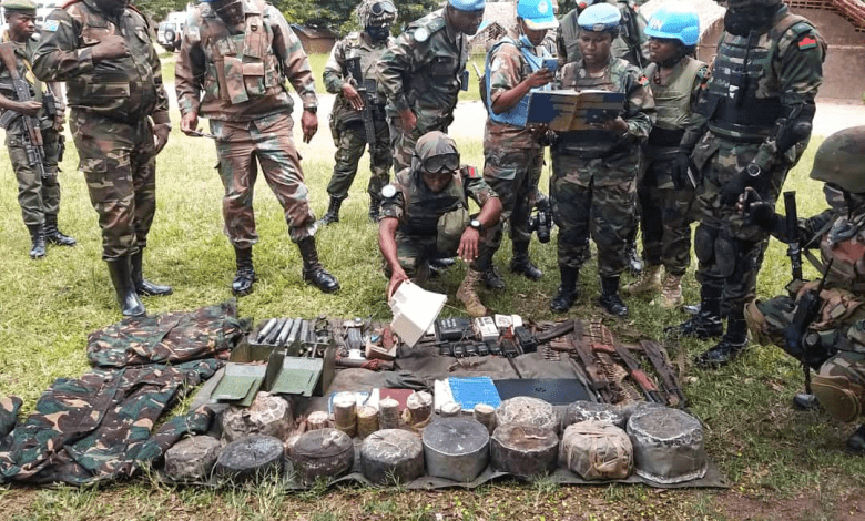 Soldiers in camouflage and UN helmets examine items laid out on the ground, discussing and taking notes.