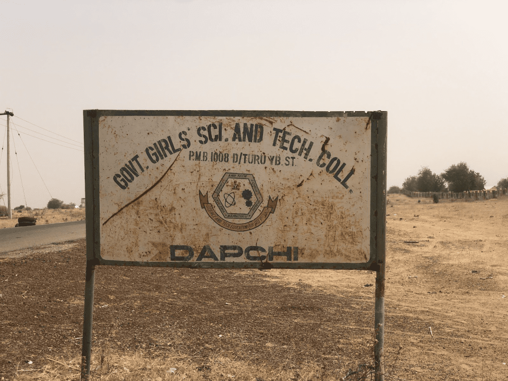 Rusty sign reading "Govt Girls Sci. and Tech. Coll. Dapchi" in a dry, rural landscape.