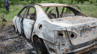 Charred remains of a burnt-out car in a grassy area with a person in patterned attire walking away in the background.