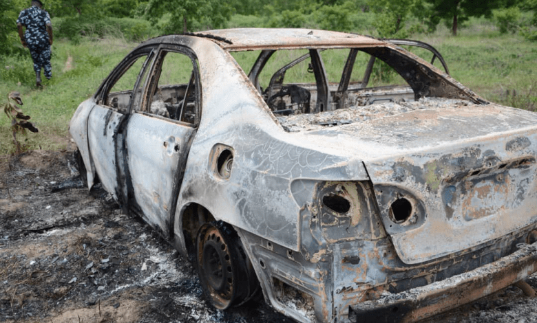 Charred remains of a burnt-out car in a grassy area with a person in patterned attire walking away in the background.