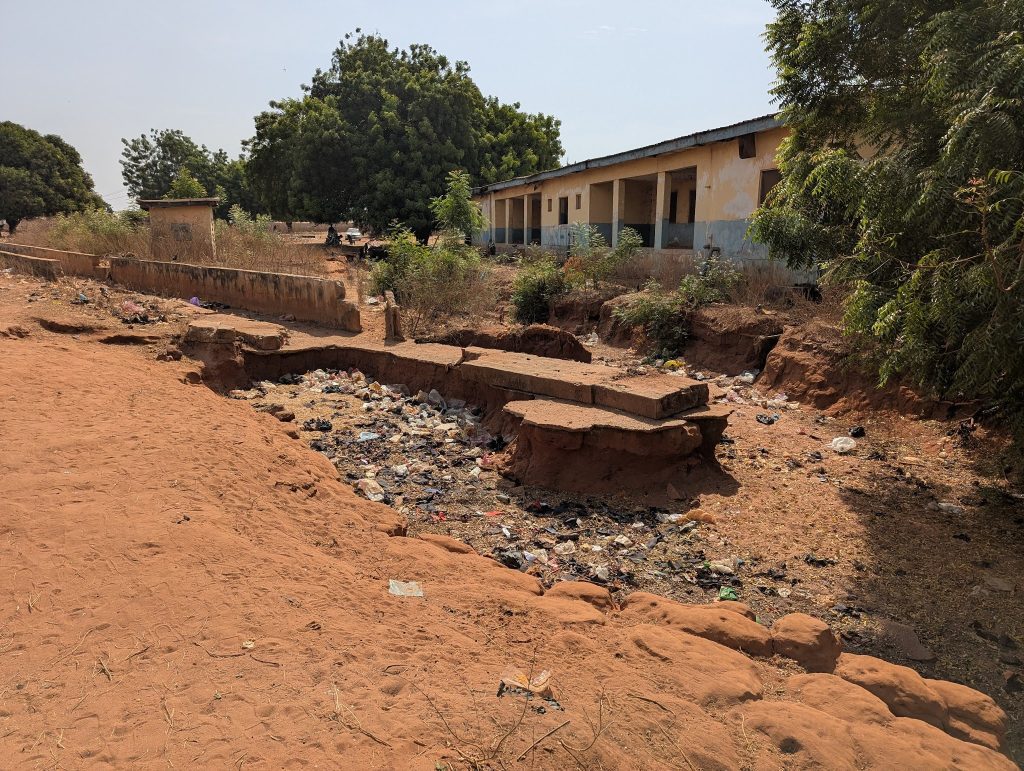 Dusty landscape with a bridge over a garbage-filled ditch, flanked by trees and a weathered building.