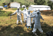 People in full protective suits, masks, and goggles standing outside near tents and a building in a rural area.