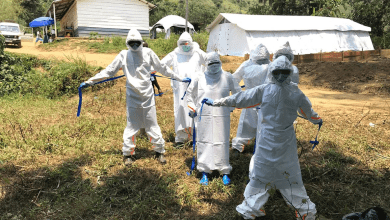 People in full protective suits, masks, and goggles standing outside near tents and a building in a rural area.
