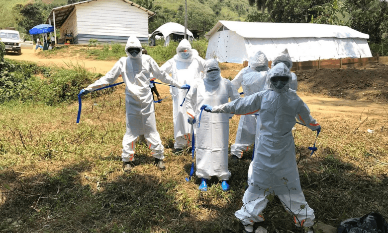 People in full protective suits, masks, and goggles standing outside near tents and a building in a rural area.