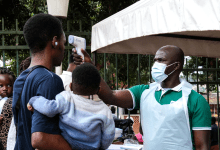 A person having their temperature checked with an infrared thermometer by a masked healthcare worker. A child is held by the person.