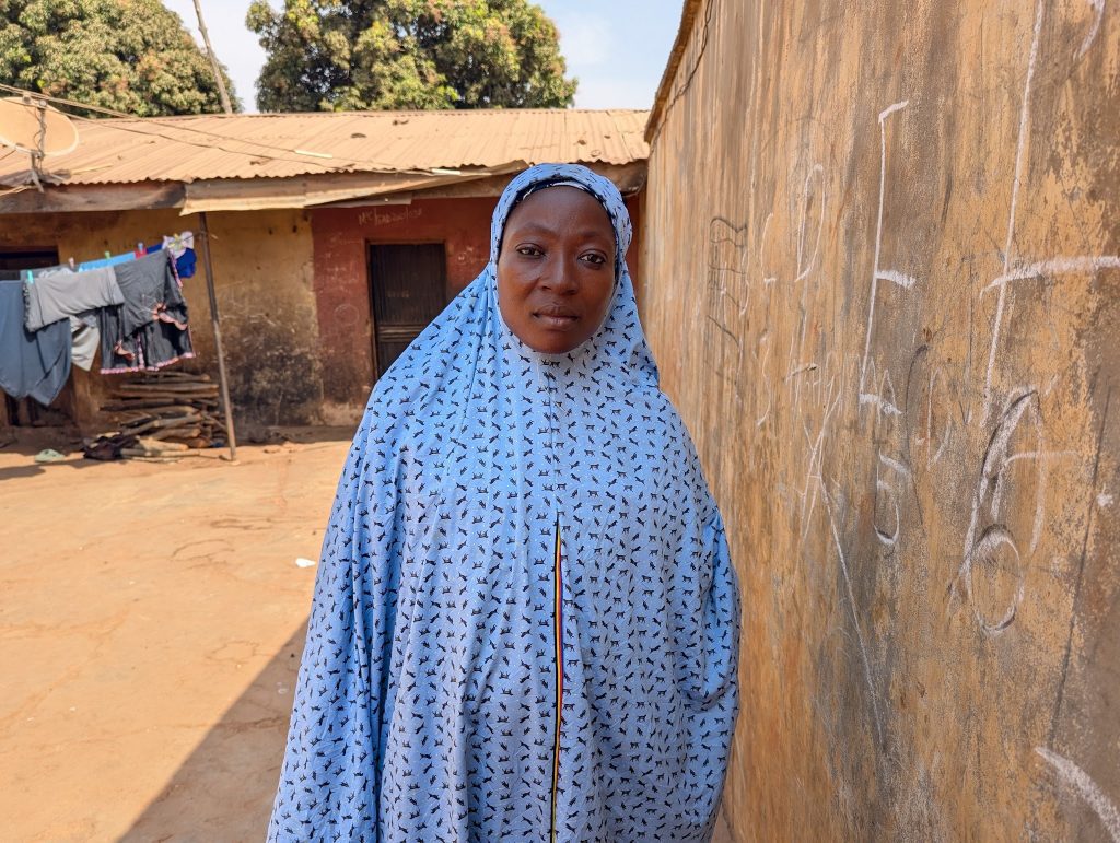 Woman in a light blue patterned hijab stands beside a wall with chalk markings, with laundry hanging in the background.