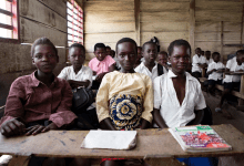 A group of children in a classroom, seated at wooden desks, looking toward the camera with notebooks in front.