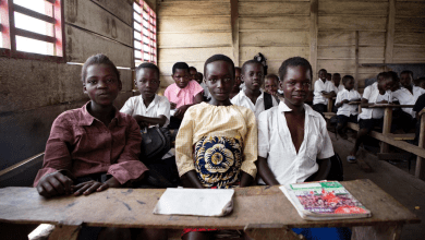 A group of children in a classroom, seated at wooden desks, looking toward the camera with notebooks in front.