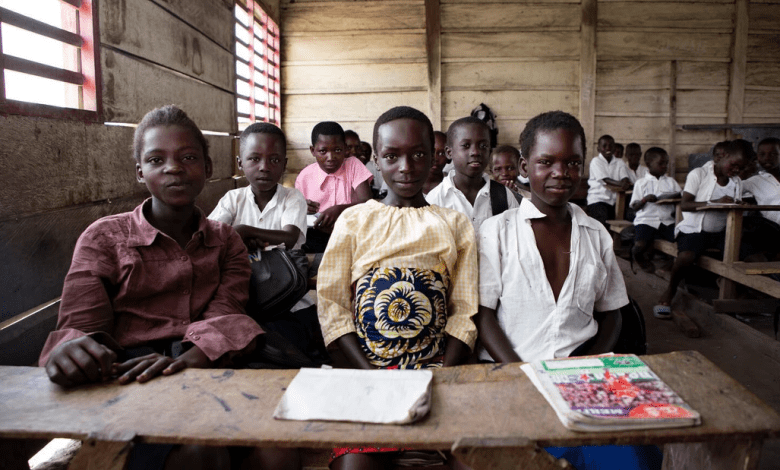 A group of children in a classroom, seated at wooden desks, looking toward the camera with notebooks in front.