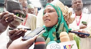 A woman in traditional attire speaks to a group of journalists holding microphones and recording devices.