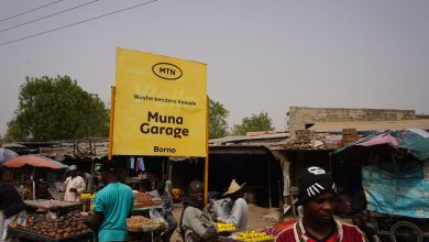 Outdoor market with a yellow "Muna Garage" sign in Borno. Vendors sell produce under makeshift stalls. People are browsing the market.