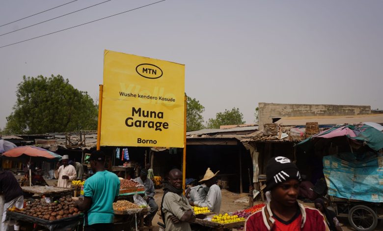 Outdoor market with a yellow "Muna Garage" sign in Borno. Vendors sell produce under makeshift stalls. People are browsing the market.