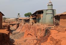 Eroded red soil surrounds a small mosque; houses and a palm tree stand nearby under a clear sky.