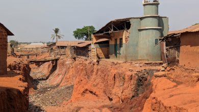 Eroded red soil surrounds a small mosque; houses and a palm tree stand nearby under a clear sky.