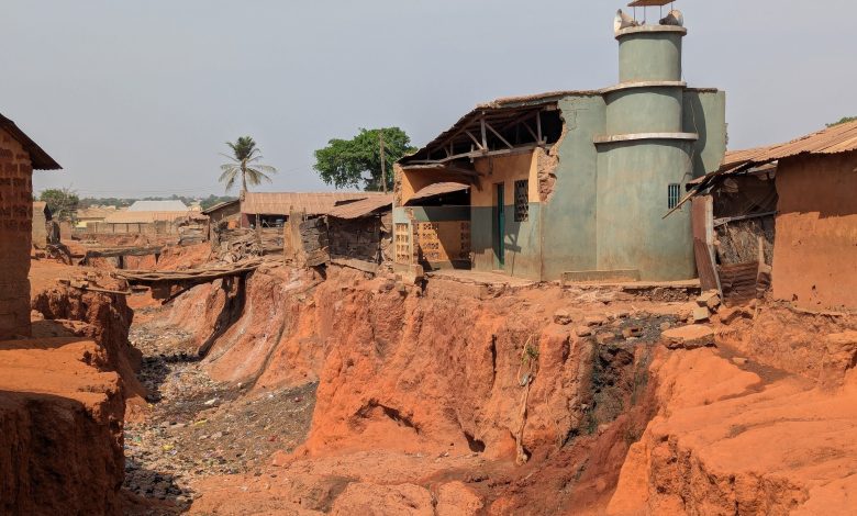 Eroded red soil surrounds a small mosque; houses and a palm tree stand nearby under a clear sky.