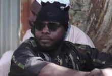 A person wearing sunglasses and a head covering sits outdoors with arms crossed, against a tree and a wall in the background.