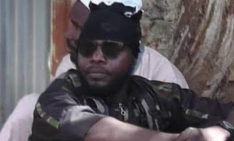 A person wearing sunglasses and a head covering sits outdoors with arms crossed, against a tree and a wall in the background.