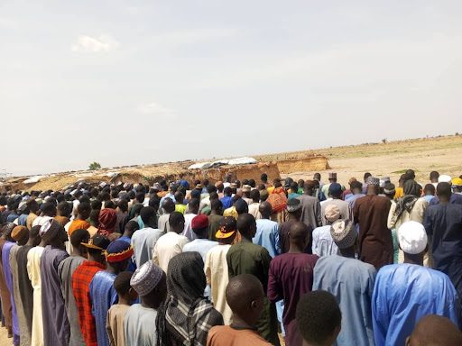 A large group of people stand in rows outdoors, appearing to observe or participate in an event under a clear sky.