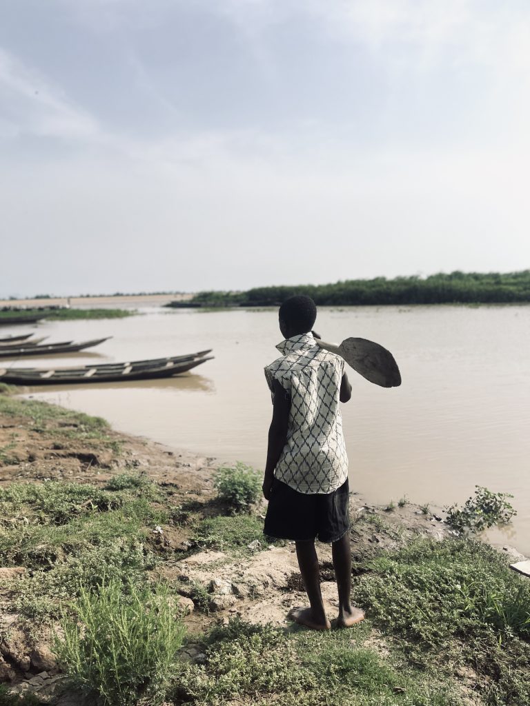 Person with a shovel looks at boats on a riverbank, surrounded by greenery and a cloudy sky.