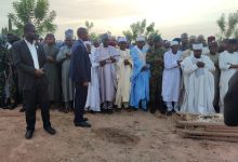 A group of people gather outdoors in traditional attire, some praying, near a covered body on a stretcher.