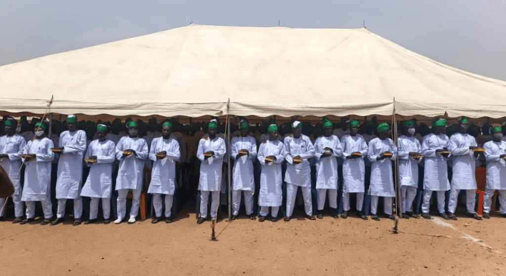 A group of men in white outfits and green hats stand in formation under a large tent outdoors.