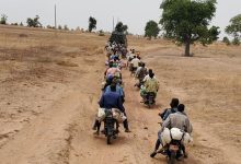 People on motorcycles follow a military vehicle on a dirt road through a dry landscape with sparse trees.