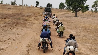 People on motorcycles follow a military vehicle on a dirt road through a dry landscape with sparse trees.